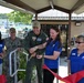A ribbon-cutting ceremony is taking place at what appears to be a military installation, likely a pool or recreational facility. A military officer in a green flight suit and a woman in a blue MWR (Morale, Welfare, and Recreation) polo shirt are jointly cutting a red ribbon with golden scissors. Several other attendees, including a Navy sailor in camouflage uniform and additional MWR staff members, are present and smiling.