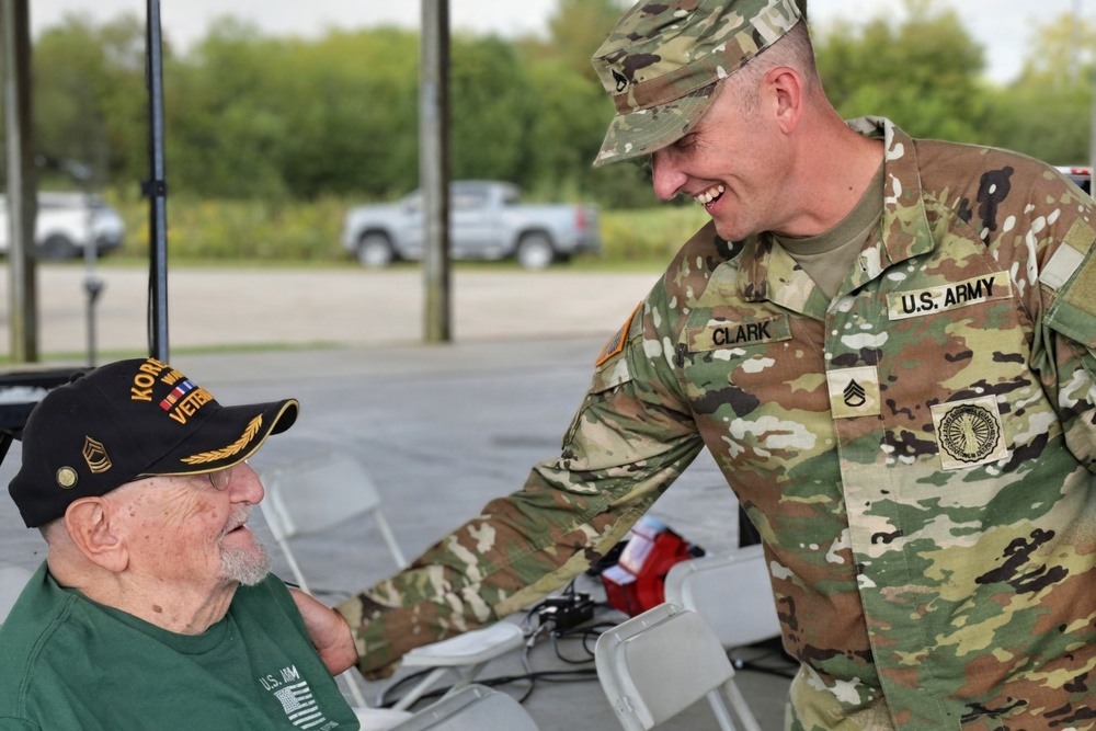 A U.S. Army soldier named Clark, wearing OCP (Operational Camouflage Pattern) uniform with a Staff Sergeant rank insignia, is smiling and leaning toward an elderly Korean War veteran seated in a chair. The veteran is wearing a black cap identifying him as a Korean War veteran and a green shirt with U.S. Army insignia. The interaction appears warm and respectful, likely at a veterans' event or ceremony held outdoors.