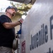 A middle-aged man wearing a black t-shirt, gray cap, and work gloves is leaning over or operating equipment next to a large tank or container labeled 'Juvenile Fi[sh]'. The setting appears to be an outdoor fish hatchery or juvenile fish facility. The image is a close-up, candid-style photograph taken in natural daylight.