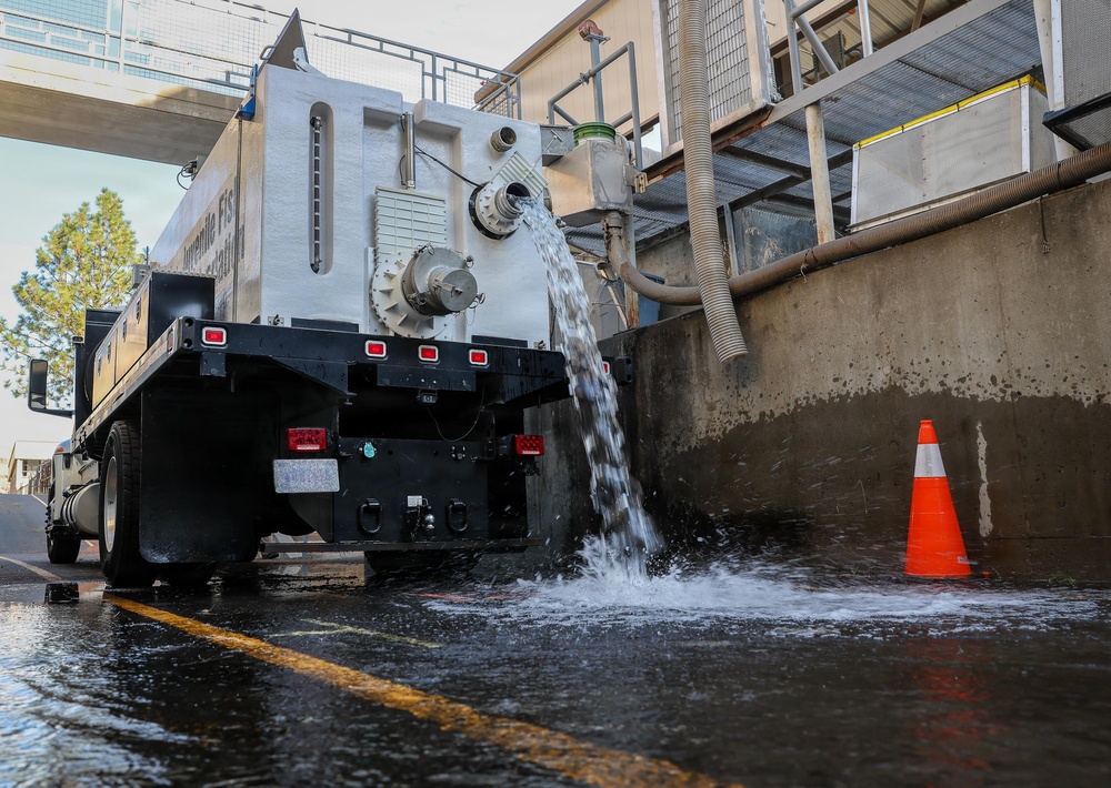 A large white tank truck is discharging water through a rear outlet pipe onto a paved surface adjacent to a concrete structure. The water is flowing forcefully from the truck's tank, pooling on the ground near an orange traffic cone. The setting appears to be an industrial or municipal facility, possibly a fish hatchery based on partial text visible on the tank.