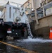 A large white tank truck is discharging water through a rear outlet pipe onto a paved surface adjacent to a concrete structure. The water is flowing forcefully from the truck's tank, pooling on the ground near an orange traffic cone. The setting appears to be an industrial or municipal facility, possibly a fish hatchery based on partial text visible on the tank.