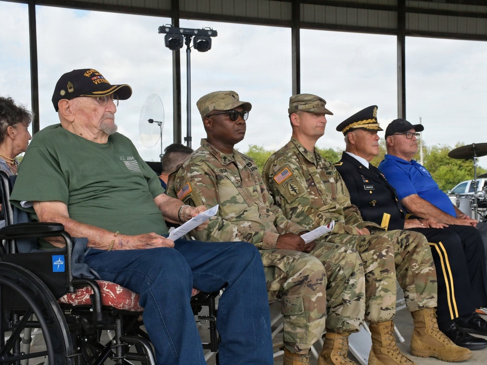 A military ceremony or commemorative event featuring a group of seated attendees including active-duty U.S. Army soldiers in OCP (Operational Camouflage Pattern) uniforms, a senior officer in dress uniform, and an elderly veteran in a wheelchair wearing a 'Korean War Veteran' cap. The gathering appears to be held under an open-air pavilion or covered outdoor venue. The mix of active military personnel and veterans suggests a formal recognition or memorial event.