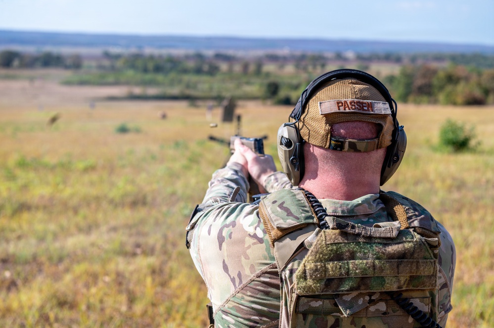A military or law enforcement personnel is seen from behind, aiming a pistol downrange at an outdoor shooting range. The individual is wearing multicam camouflage uniform, a plate carrier vest, tactical hearing protection, and eye protection. A name tape reading 'PASSEN' is visible on the back of their cap.