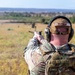 A military or law enforcement personnel is seen from behind, aiming a pistol downrange at an outdoor shooting range. The individual is wearing multicam camouflage uniform, a plate carrier vest, tactical hearing protection, and eye protection. A name tape reading 'PASSEN' is visible on the back of their cap.