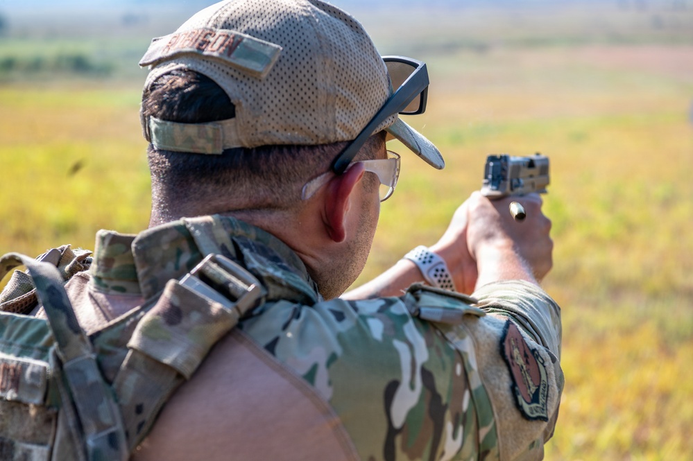 A military or law enforcement personnel is seen from behind, firing a semi-automatic pistol at an outdoor range. The individual is wearing MultiCam/OCP camouflage uniform with a tactical vest, a tan baseball cap, protective eyewear, and hearing protection. A spent brass casing is visible in the air near the firearm, indicating an active firing moment.