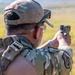 A military or law enforcement personnel is seen from behind, firing a semi-automatic pistol at an outdoor range. The individual is wearing MultiCam/OCP camouflage uniform with a tactical vest, a tan baseball cap, protective eyewear, and hearing protection. A spent brass casing is visible in the air near the firearm, indicating an active firing moment.