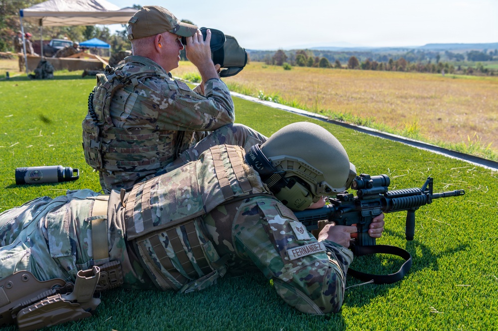 Two military personnel in OCP (Operational Camouflage Pattern) uniforms are conducting a shooting range exercise. One soldier, identified by name tape as 'FERNANDEZ,' is in the prone position aiming an M4/M16-style rifle downrange, while a second soldier sits behind them using binoculars to observe the target area. The setting appears to be an outdoor military firing range with a green grass surface and open terrain in the background.