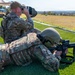 Two military personnel in OCP (Operational Camouflage Pattern) uniforms are conducting a shooting range exercise. One soldier, identified by name tape as 'FERNANDEZ,' is in the prone position aiming an M4/M16-style rifle downrange, while a second soldier sits behind them using binoculars to observe the target area. The setting appears to be an outdoor military firing range with a green grass surface and open terrain in the background.