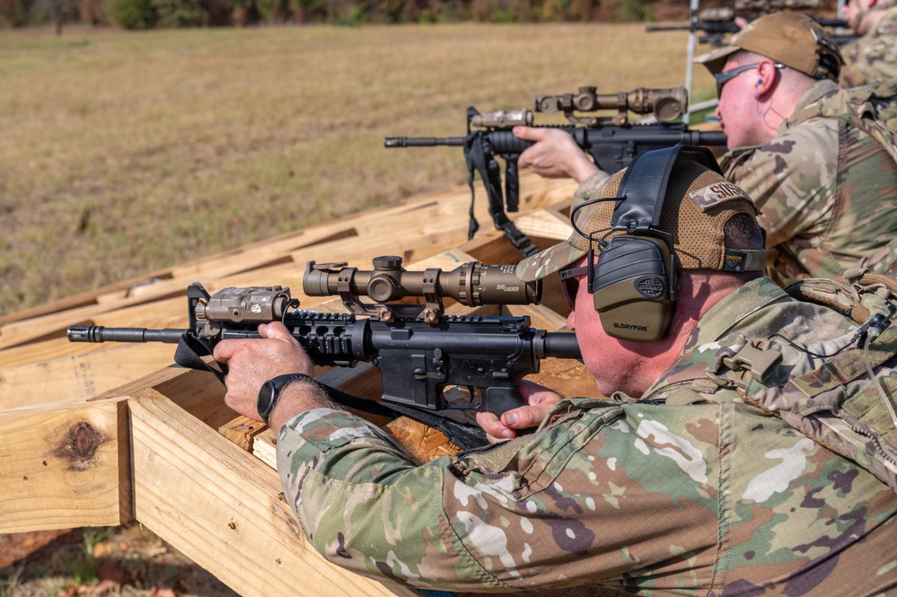 Two individuals in military camouflage (MultiCam/OCP pattern) are positioned at a wooden shooting bench at an outdoor firing range. Both are aiming rifles equipped with optics and accessories. The setting appears to be a formal military or law enforcement training exercise.