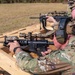 Two individuals in military camouflage (MultiCam/OCP pattern) are positioned at a wooden shooting bench at an outdoor firing range. Both are aiming rifles equipped with optics and accessories. The setting appears to be a formal military or law enforcement training exercise.