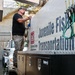 A worker is operating equipment on a US Army Corps of Engineers 'Juvenile Fish Transportation' truck or tanker vehicle. The man is standing on the side of the large specialized transport unit, appearing to manage connections or controls. The setting appears to be a fish facility or dam infrastructure site.