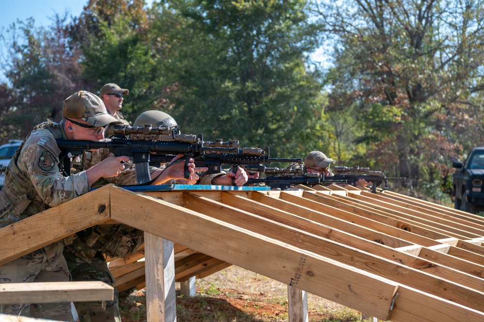 Multiple personnel in military/tactical camouflage uniforms are engaged in a rifle shooting exercise at an outdoor firing range. They are positioned behind a wooden bench rest structure, aiming rifles equipped with optics and accessories downrange. A supervisor or range safety officer stands behind the line observing the exercise.