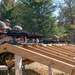 Multiple personnel in military/tactical camouflage uniforms are engaged in a rifle shooting exercise at an outdoor firing range. They are positioned behind a wooden bench rest structure, aiming rifles equipped with optics and accessories downrange. A supervisor or range safety officer stands behind the line observing the exercise.
