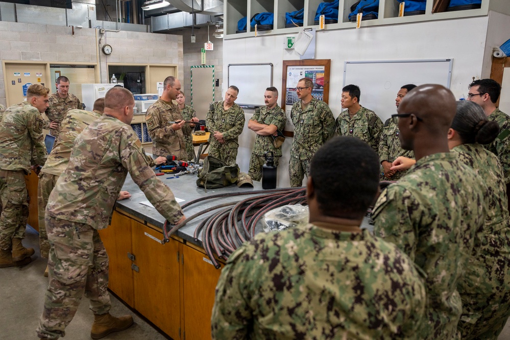A joint military training session is taking place in what appears to be a technical workshop or maintenance facility. Personnel wearing both Army Combat Uniforms (ACU/OCP pattern) and Navy Working Uniforms (NWU Type III) are gathered around a workbench where an instructor is demonstrating equipment. The group appears to be receiving hands-on instruction involving cables, hoses, and various tools.