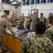 A joint military training session is taking place in what appears to be a technical workshop or maintenance facility. Personnel wearing both Army Combat Uniforms (ACU/OCP pattern) and Navy Working Uniforms (NWU Type III) are gathered around a workbench where an instructor is demonstrating equipment. The group appears to be receiving hands-on instruction involving cables, hoses, and various tools.