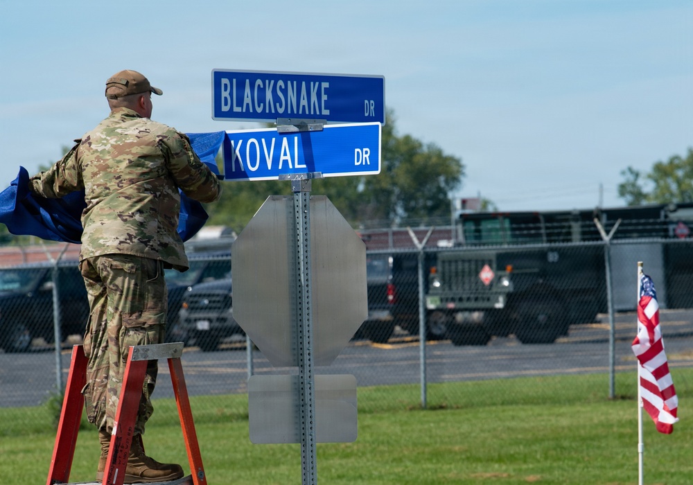A military service member in camouflage uniform stands on a red ladder while unveiling or installing street signs at an intersection labeled 'Blacksnake Dr' and 'Koval Dr' on what appears to be a military installation. A blue covering is being removed from the signs, suggesting a dedication or naming ceremony. An American flag on a small pole is visible in the background near a fenced vehicle lot.