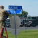 A military service member in camouflage uniform stands on a red ladder while unveiling or installing street signs at an intersection labeled 'Blacksnake Dr' and 'Koval Dr' on what appears to be a military installation. A blue covering is being removed from the signs, suggesting a dedication or naming ceremony. An American flag on a small pole is visible in the background near a fenced vehicle lot.