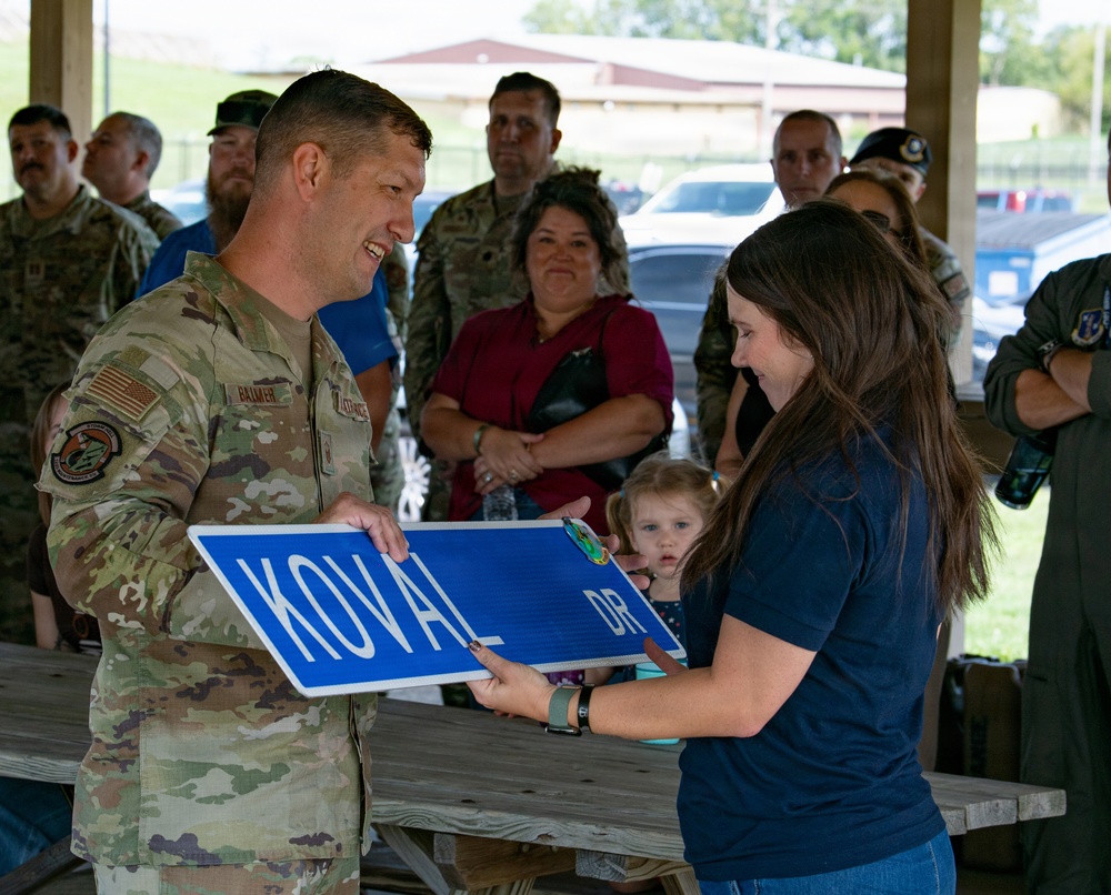 A military ceremony appears to be taking place under an outdoor pavilion, where a uniformed service member is presenting a street sign reading 'KOVAL DR' to a civilian woman. The event appears to be a street dedication or naming ceremony, likely honoring someone named Koval. A crowd of military personnel and civilians, including a small child, are gathered as witnesses in the background.