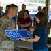 A military ceremony appears to be taking place under an outdoor pavilion, where a uniformed service member is presenting a street sign reading 'KOVAL DR' to a civilian woman. The event appears to be a street dedication or naming ceremony, likely honoring someone named Koval. A crowd of military personnel and civilians, including a small child, are gathered as witnesses in the background.