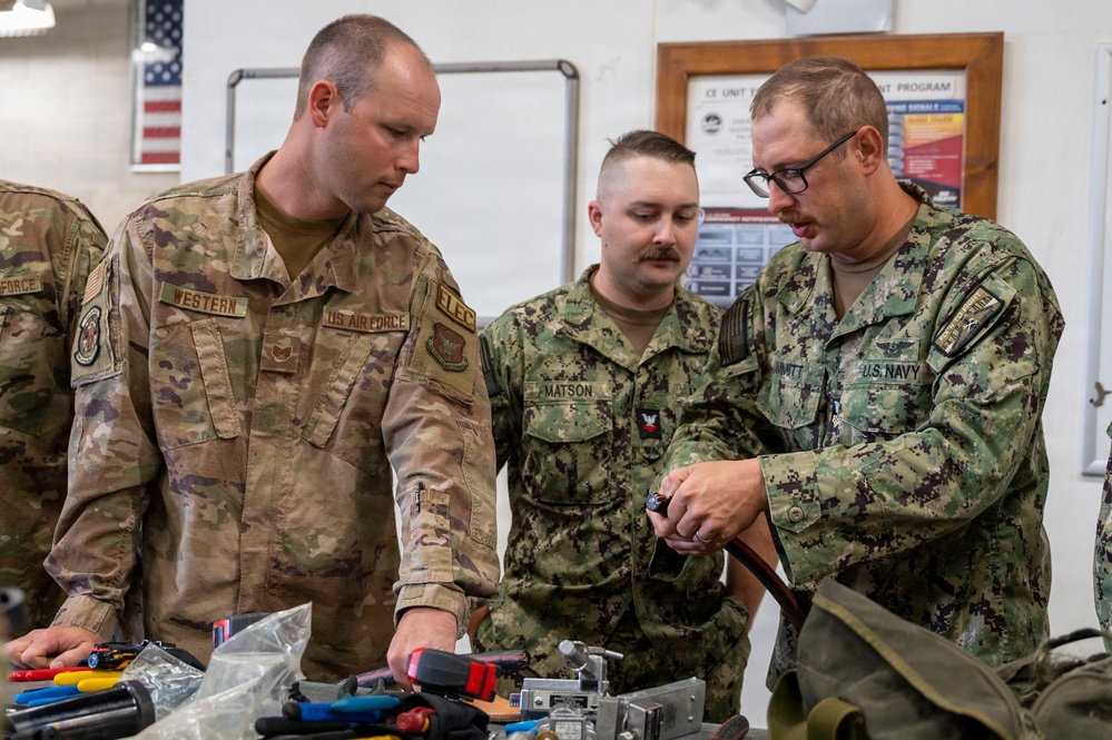 Three military personnel from different branches — U.S. Air Force and U.S. Navy — are gathered around a workbench examining tools and equipment. The individuals are identifiable by their name tapes and branch insignia, with 'WESTERN' and 'MATSON' visible, along with a U.S. Navy member wearing glasses. The setting appears to be an indoor military workshop or training facility.