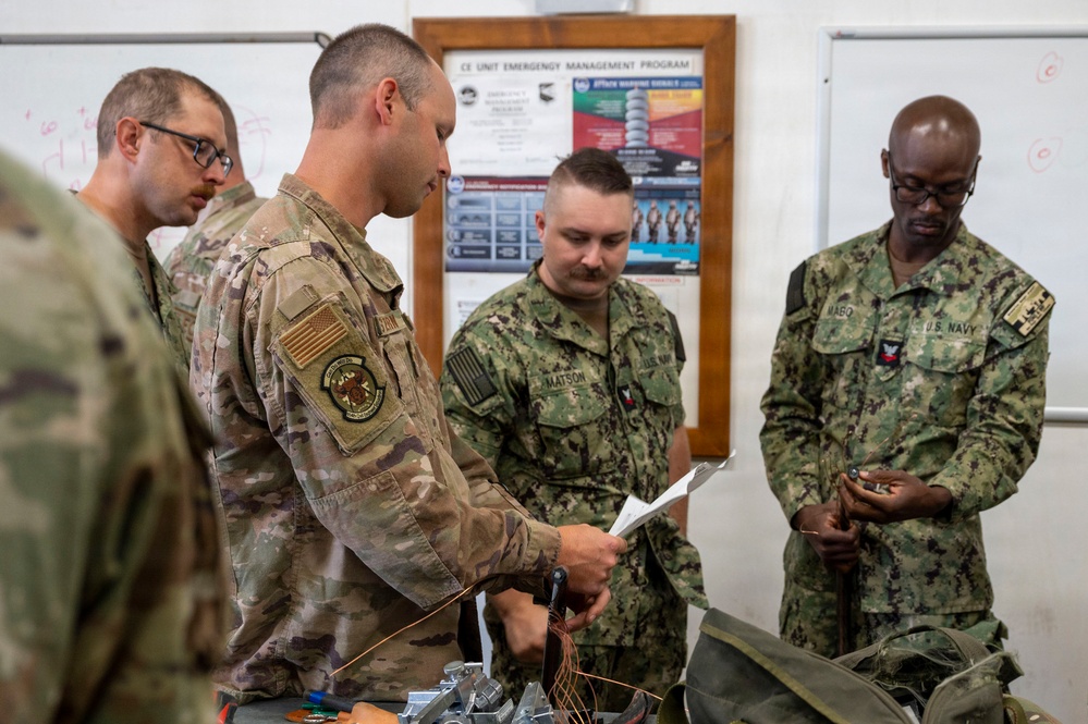 Military personnel from multiple branches are gathered around a table with equipment in what appears to be a training or briefing room. The group includes U.S. Air Force and U.S. Navy members examining materials and documents. A bulletin board in the background displays a 'CE Unit Emergency Management Program' sign.