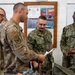 Military personnel from multiple branches are gathered around a table with equipment in what appears to be a training or briefing room. The group includes U.S. Air Force and U.S. Navy members examining materials and documents. A bulletin board in the background displays a 'CE Unit Emergency Management Program' sign.
