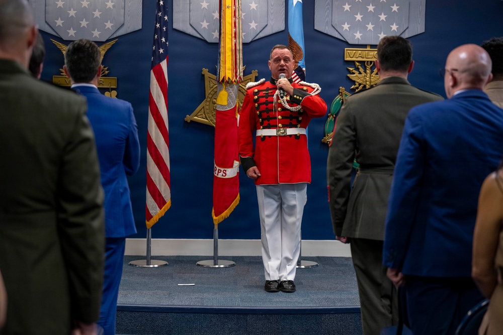 A man in a distinctive red ceremonial military band uniform is speaking into a microphone on a raised platform, surrounded by flags including the United States flag and what appears to be a Marine Corps flag. The setting appears to be an official military ceremony or event, with attendees in military uniforms and civilian suits standing on either side. The backdrop features a blue wall with military insignia, stars, and the word 'VALOR'.