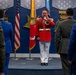 A man in a distinctive red ceremonial military band uniform is speaking into a microphone on a raised platform, surrounded by flags including the United States flag and what appears to be a Marine Corps flag. The setting appears to be an official military ceremony or event, with attendees in military uniforms and civilian suits standing on either side. The backdrop features a blue wall with military insignia, stars, and the word 'VALOR'.