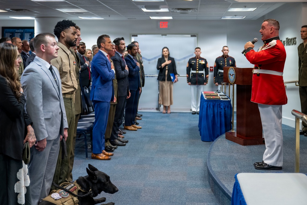 A formal military ceremony is taking place indoors, likely at the Pentagon based on the Department of Defense seal visible on the podium. A Marine officer in dress red uniform is speaking at a podium while attendees — including military personnel in dress uniforms and civilians in business attire — stand with hands over hearts, suggesting the playing or singing of the national anthem. A black service dog wearing a vest is lying on the floor near the front row.