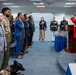 A formal military ceremony is taking place indoors, likely at the Pentagon based on the Department of Defense seal visible on the podium. A Marine officer in dress red uniform is speaking at a podium while attendees — including military personnel in dress uniforms and civilians in business attire — stand with hands over hearts, suggesting the playing or singing of the national anthem. A black service dog wearing a vest is lying on the floor near the front row.