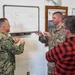 A group of military personnel and a civilian are engaged in a technical discussion in front of a whiteboard containing electrical circuit diagrams. The group includes members from the U.S. Navy (digital camouflage uniforms) and U.S. Air Force (OCP/multicam uniforms), along with one civilian in a red flannel shirt. The setting appears to be a military facility room, with a 'CE Unit Emergency Management Program' board visible on the wall.