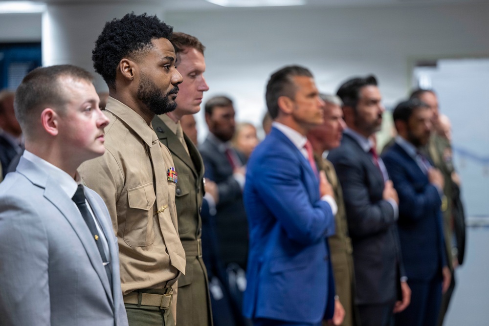 A group of individuals standing in a line, appearing to be at a formal ceremony or official event. One person in the foreground is wearing a U.S. Marine Corps dress uniform with visible ribbons and insignia, while others are dressed in civilian business attire. Several individuals in the background have their hands placed over their hearts, suggesting a pledge or national anthem moment.