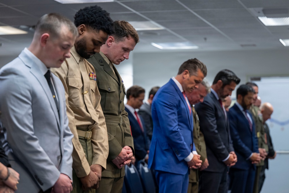 A group of men standing in a line with heads bowed in what appears to be a moment of prayer or silence in an indoor office or conference room setting. The group includes individuals in military uniforms and civilian business attire. The solemn posture and clasped hands suggest a formal ceremonial or commemorative occasion.