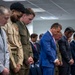 A group of men standing in a line with heads bowed in what appears to be a moment of prayer or silence in an indoor office or conference room setting. The group includes individuals in military uniforms and civilian business attire. The solemn posture and clasped hands suggest a formal ceremonial or commemorative occasion.