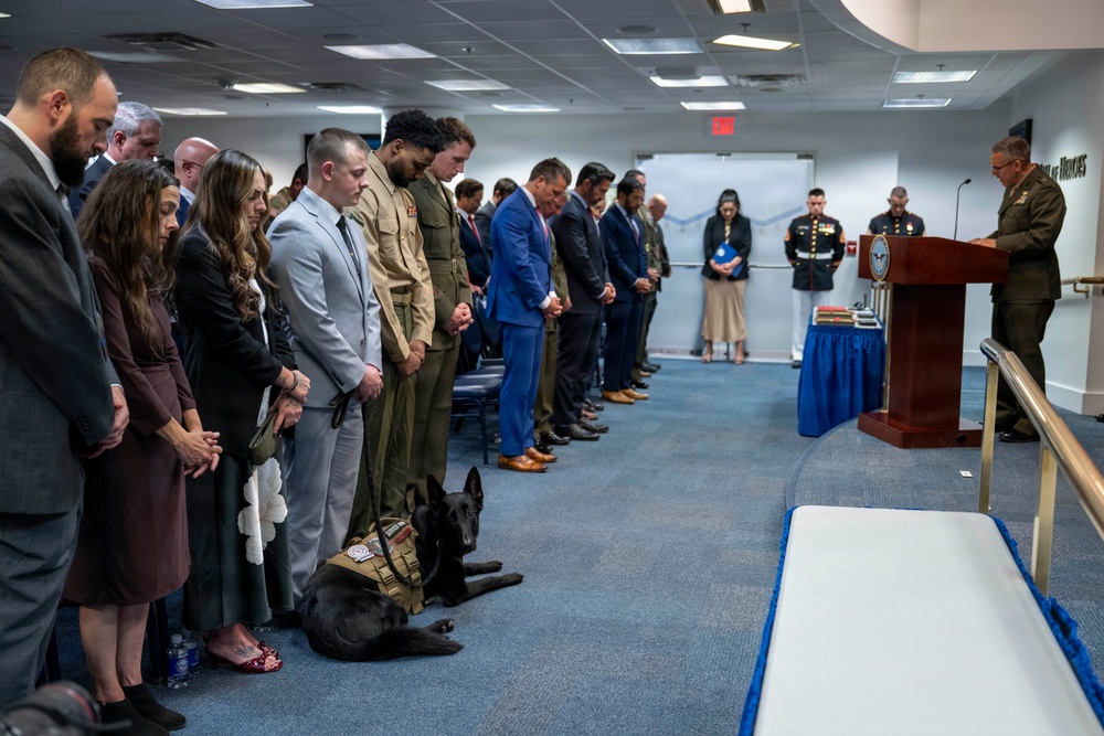 A formal military ceremony is taking place in an official government room, with a group of attendees standing in a line with heads bowed in prayer or moment of silence. A military officer in dress uniform stands at a wooden podium delivering remarks. A black military working dog wearing a tactical vest lies calmly on the floor among the attendees.