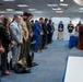 A formal military ceremony is taking place in an official government room, with a group of attendees standing in a line with heads bowed in prayer or moment of silence. A military officer in dress uniform stands at a wooden podium delivering remarks. A black military working dog wearing a tactical vest lies calmly on the floor among the attendees.