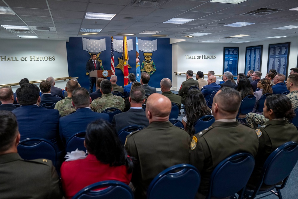 A formal ceremony or presentation is taking place in the Pentagon's Hall of Heroes, a room dedicated to Medal of Honor recipients. A speaker in civilian attire addresses an audience of mixed military personnel in various uniforms and civilians seated in blue chairs. The backdrop features large Medal of Honor insignia displayed on a blue wall flanked by American and military service flags.