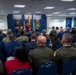 A formal ceremony or presentation is taking place in the Pentagon's Hall of Heroes, a room dedicated to Medal of Honor recipients. A speaker in civilian attire addresses an audience of mixed military personnel in various uniforms and civilians seated in blue chairs. The backdrop features large Medal of Honor insignia displayed on a blue wall flanked by American and military service flags.
