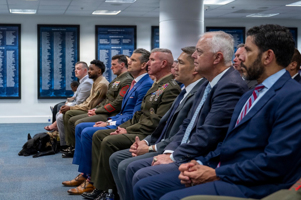 A formal gathering showing a row of seated individuals including military personnel in uniform and civilians in business attire, likely attending an official ceremony or briefing. The setting appears to be a government or military facility, with framed honor boards or name plaques visible on the walls in the background. A military working dog is also visible at the far left of the image.