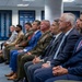 A formal gathering showing a row of seated individuals including military personnel in uniform and civilians in business attire, likely attending an official ceremony or briefing. The setting appears to be a government or military facility, with framed honor boards or name plaques visible on the walls in the background. A military working dog is also visible at the far left of the image.