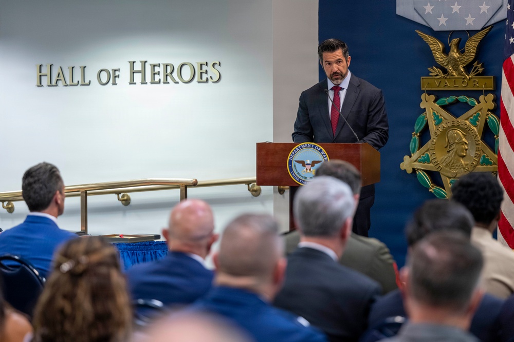 A man in a dark suit with a red tie is speaking at a podium bearing the Department of War seal in the Pentagon's Hall of Heroes. Behind him is a large Medal of Honor display with the word 'VALOR' and an American flag. An audience of military and civilian personnel is seated facing the speaker.