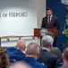 A man in a dark suit with a red tie is speaking at a podium bearing the Department of War seal in the Pentagon's Hall of Heroes. Behind him is a large Medal of Honor display with the word 'VALOR' and an American flag. An audience of military and civilian personnel is seated facing the speaker.