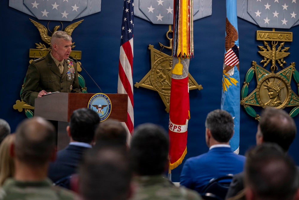 A senior U.S. Marine Corps officer in dress uniform is delivering remarks at a podium bearing the Department of Defense seal inside what appears to be the Pentagon. The backdrop features large decorative Medal of Honor replicas, flags including the U.S. flag and a Marine Corps flag, and gold emblems with the word 'VALOR'. An audience of military personnel and civilians is seated facing the speaker.