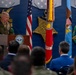 A senior U.S. Marine Corps officer in dress uniform is delivering remarks at a podium bearing the Department of Defense seal inside what appears to be the Pentagon. The backdrop features large decorative Medal of Honor replicas, flags including the U.S. flag and a Marine Corps flag, and gold emblems with the word 'VALOR'. An audience of military personnel and civilians is seated facing the speaker.