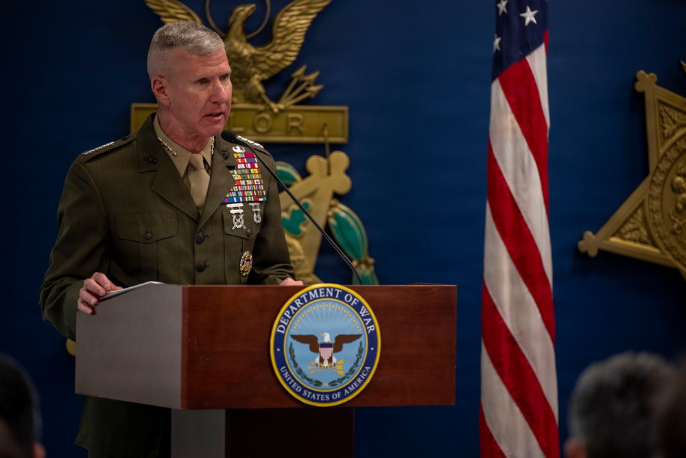 A senior U.S. Marine Corps officer in dress uniform is speaking at a podium bearing a seal that reads 'Department of War, United States of America.' The setting appears to be an official government or military venue, with a U.S. flag and decorative gold eagle emblem visible in the background. The officer displays numerous service ribbons and insignia consistent with a high-ranking general officer.