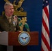 A senior U.S. Marine Corps officer in dress uniform is speaking at a podium bearing a seal that reads 'Department of War, United States of America.' The setting appears to be an official government or military venue, with a U.S. flag and decorative gold eagle emblem visible in the background. The officer displays numerous service ribbons and insignia consistent with a high-ranking general officer.