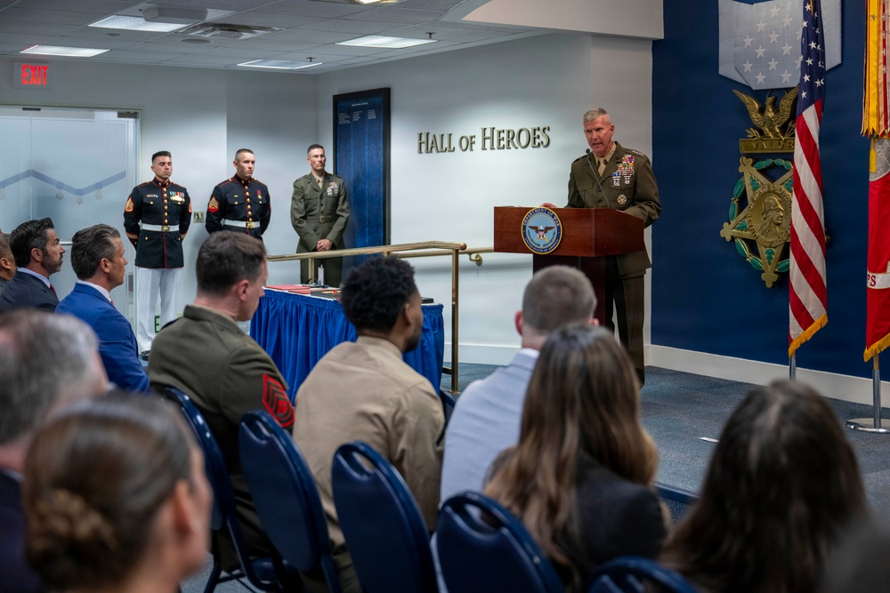 A military ceremony is taking place in the Pentagon's Hall of Heroes, where a senior military officer in Marine Corps dress uniform is delivering remarks at a podium bearing the Department of Defense seal. Several Marines in dress blues and service uniforms stand at attention in the background, while an audience of military personnel and civilians is seated in the foreground. The Medal of Honor display and American flag are prominently visible on the right side of the room.