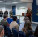 A military ceremony is taking place in the Pentagon's Hall of Heroes, where a senior military officer in Marine Corps dress uniform is delivering remarks at a podium bearing the Department of Defense seal. Several Marines in dress blues and service uniforms stand at attention in the background, while an audience of military personnel and civilians is seated in the foreground. The Medal of Honor display and American flag are prominently visible on the right side of the room.