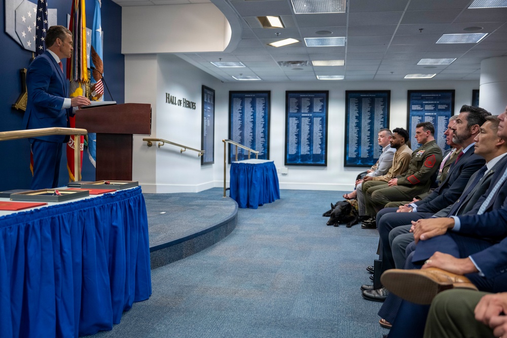 A formal ceremony is taking place in what appears to be the Pentagon's Hall of Heroes, where a man in a blue suit is speaking at a wooden podium. An audience of mixed civilians and military personnel in uniform are seated and listening attentively. Blue-skirted tables with presentation items are visible in the foreground.