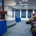 A formal ceremony is taking place in what appears to be the Pentagon's Hall of Heroes, where a man in a blue suit is speaking at a wooden podium. An audience of mixed civilians and military personnel in uniform are seated and listening attentively. Blue-skirted tables with presentation items are visible in the foreground.